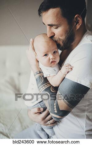 Stock Image - Caucasian father with tattoos on arms kissing baby son. Fotosearch