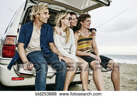 Stock Photo - Caucasian friends sitting in car hatch at beach. Fotosearch