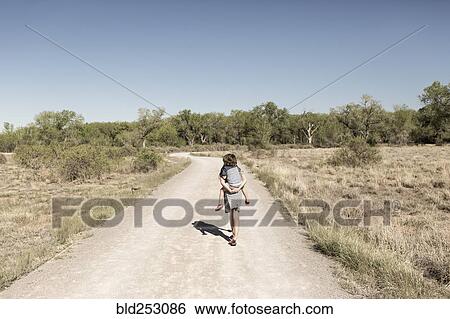Stock Photograph - Caucasian girl carrying brother piggyback on dirt path. Fotosearch