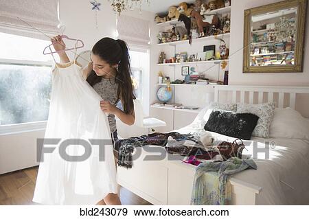 Stock Photo - Caucasian girl examining white dress in bedroom. Fotosearch
