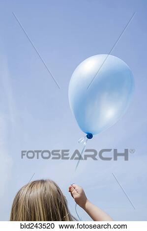Stock Image - Caucasian girl holding blue balloon in blue sky. Fotosearch