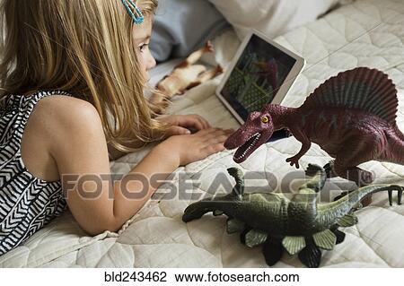 Stock Image - Caucasian girl laying on bed with toy dinosaurs using digital tablet. Fotosearch