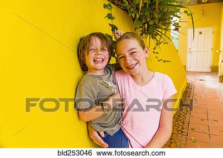 Stock Photograph - Caucasian girl leaning on yellow wall holding brother. Fotosearch