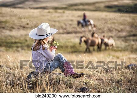 Stock Image - Caucasian girl sitting on rock in field holding flower. Fotosearch