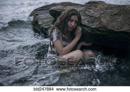 Picture - Caucasian girl sitting on rock in ocean. Fotosearch