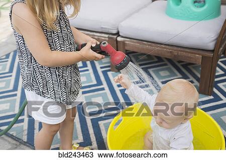 Stock Image - Caucasian girl spraying baby brother in bucket with garden hose. Fotosearch