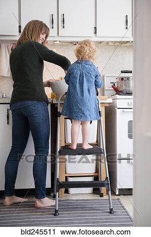 Stock Image - Caucasian girl standing on ladder cooking with mother in kitchen. Fotosearch