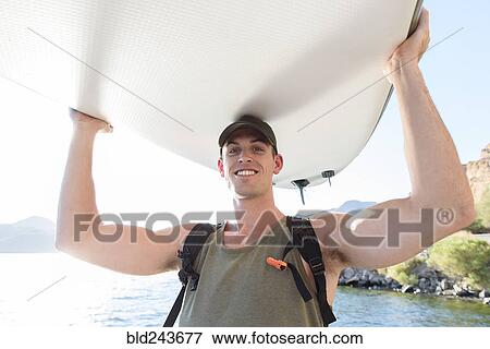 Stock Photo - Caucasian man carrying paddleboard at river. Fotosearch