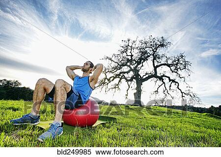 Stock Photography - Caucasian man doing sit-ups on fitness ball in field near tree. Fotosearch