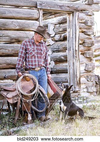 Stock Image - Caucasian man holding saddle and blanket looking at dog. Fotosearch