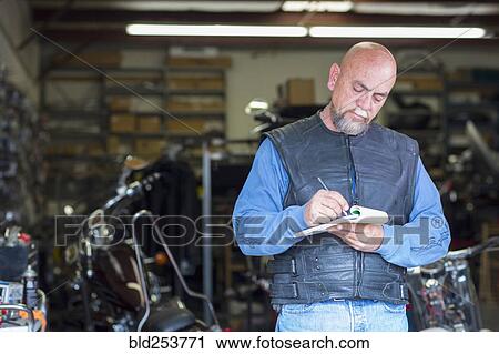Stock Image - Caucasian man near motorcycle writing on notepad. Fotosearch
