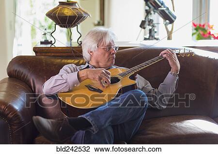 Stock Image - Caucasian man playing guitar on sofa. Fotosearch