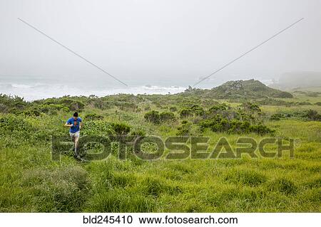 Stock Image - Caucasian man running on path near ocean beach. Fotosearch