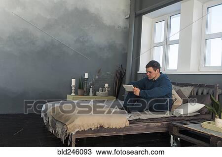 Stock Image - Caucasian man sitting in bed using digital tablet. Fotosearch