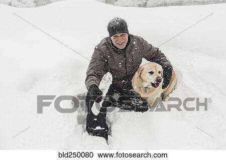 Stock Image - Caucasian man sitting in snow hugging dog. Fotosearch