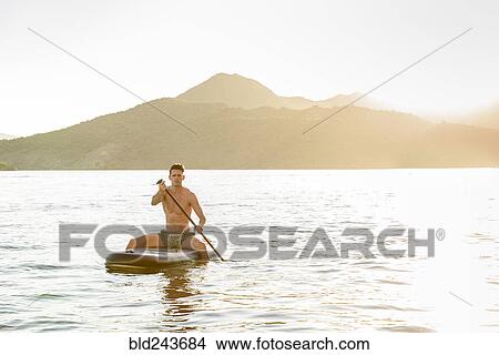 Picture - Caucasian man sitting on paddleboard in river. Fotosearch