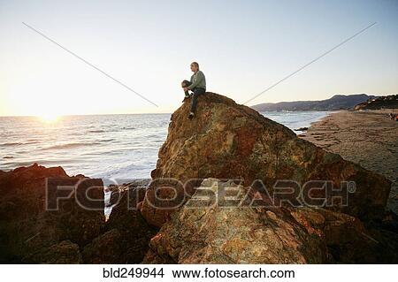 Picture - Caucasian man sitting on rock formation near ocean. Fotosearch