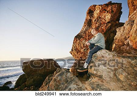 Stock Image - Caucasian man sitting on rock formation at beach. Fotosearch