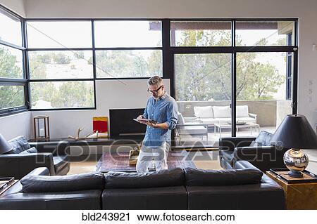Stock Image - Caucasian man standing in livingroom using digital tablet. Fotosearch