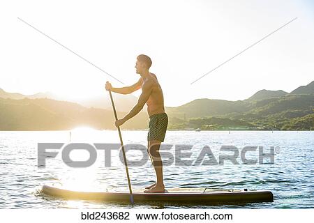 Stock Image - Caucasian man standing on paddleboard in river. Fotosearch