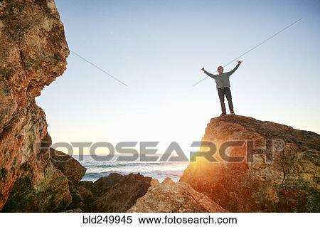 Stock Photography - Caucasian man standing with arms raised on rock at ocean. Fotosearch