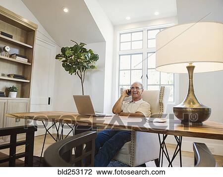 Stock Image - Caucasian man talking on cell phone in home office. Fotosearch