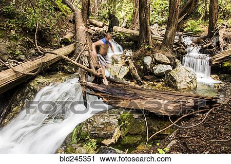 Stock Image - Caucasian man walking over waterfall on log in woods. Fotosearch