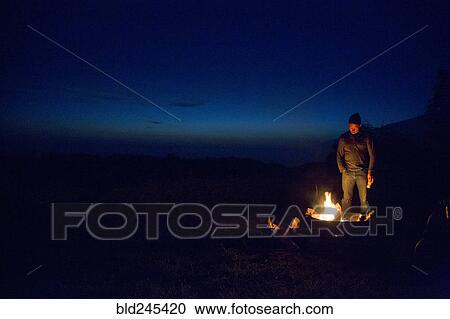 Stock Image - Caucasian man watching campfire at night. Fotosearch