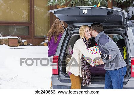 Stock Image - Caucasian man whispering to woman carrying gifts from car in winter. Fotosearch