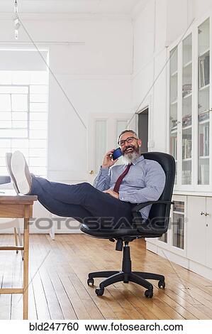 Stock Image - Caucasian man with feet up talking on cell phone. Fotosearch