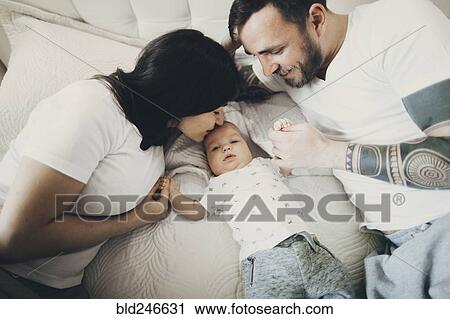 Stock Image - Caucasian mother and father holding hands with baby son on bed. Fotosearch