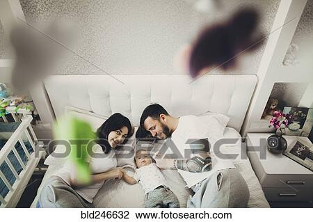 Stock Image - Caucasian mother and father holding hands with baby son on bed. Fotosearch