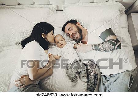 Stock Image - Caucasian mother and father holding hands with baby son on bed. Fotosearch