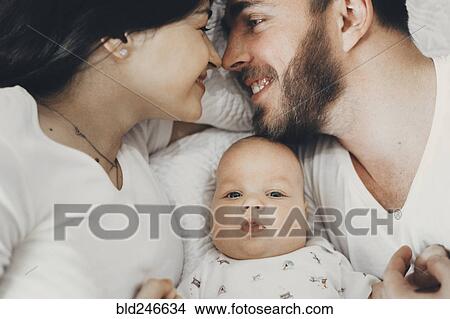 Picture - Caucasian mother and father laying on bed with baby son. Fotosearch
