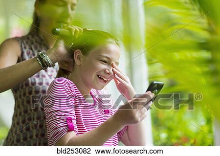 Stock Image - Caucasian mother brushing hair of daughter texting on cell phone. Fotosearch