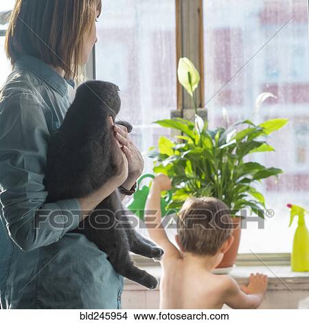 Picture - Caucasian mother holding cat watching son watering plant. Fotosearch
