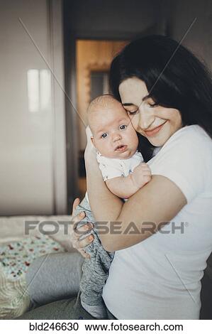 Stock Image - Caucasian mother kneeling on bed hugging baby son. Fotosearch