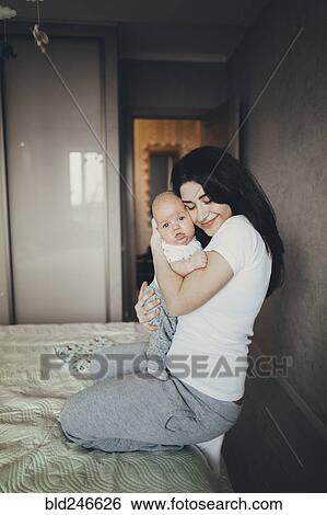 Stock Photograph - Caucasian mother kneeling on bed hugging baby son. Fotosearch