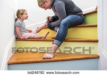 Stock Image - Caucasian mother painting toenails of daughter on multicolor staircase. Fotosearch