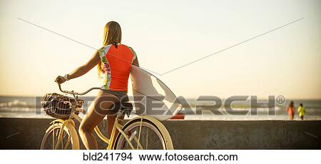 Picture - Caucasian teenage girl carrying surfboard on bicycle. Fotosearch