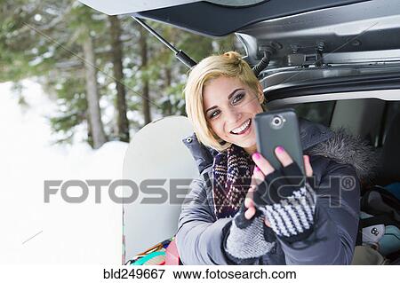 Stock Photo - Caucasian woman at car posing for cell phone selfie in winter. Fotosearch