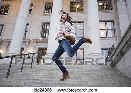 Stock Image - Caucasian woman carrying backpack jumping in snow on staircase. Fotosearch