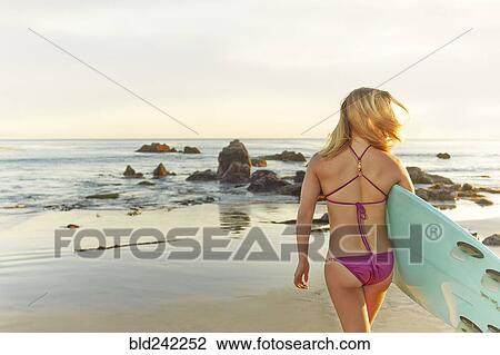 Stock Image - Caucasian woman carrying surfboard on beach. Fotosearch