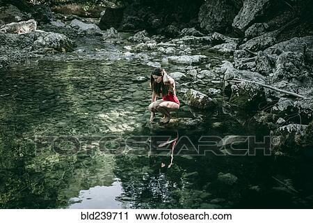 Stock Image - Caucasian woman crouching on rocks at pool of water. Fotosearch