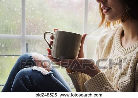 Stock Photography - Caucasian woman drinking coffee near rainy window. Fotosearch