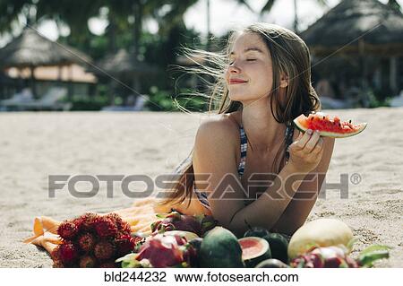 Stock Image - Caucasian woman eating watermelon on beach. Fotosearch