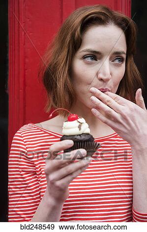 Stock Photo - Caucasian woman holding a cupcake licking finger. Fotosearch