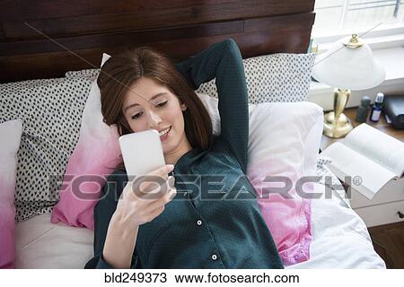 Stock Image - Caucasian woman laying in bed texting on cell phone. Fotosearch