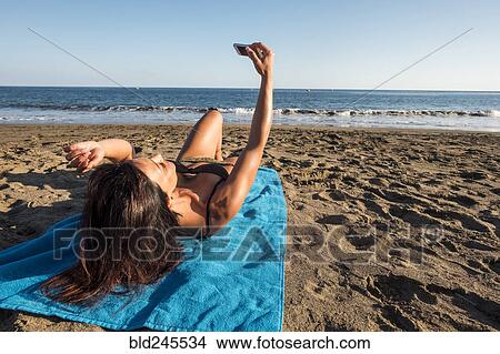 Picture - Caucasian woman laying on blanket on beach posing for cell phone selfie. Fotosearch