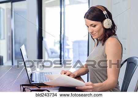 Picture - Caucasian woman listening to headphones reading paperwork using laptop. Fotosearch